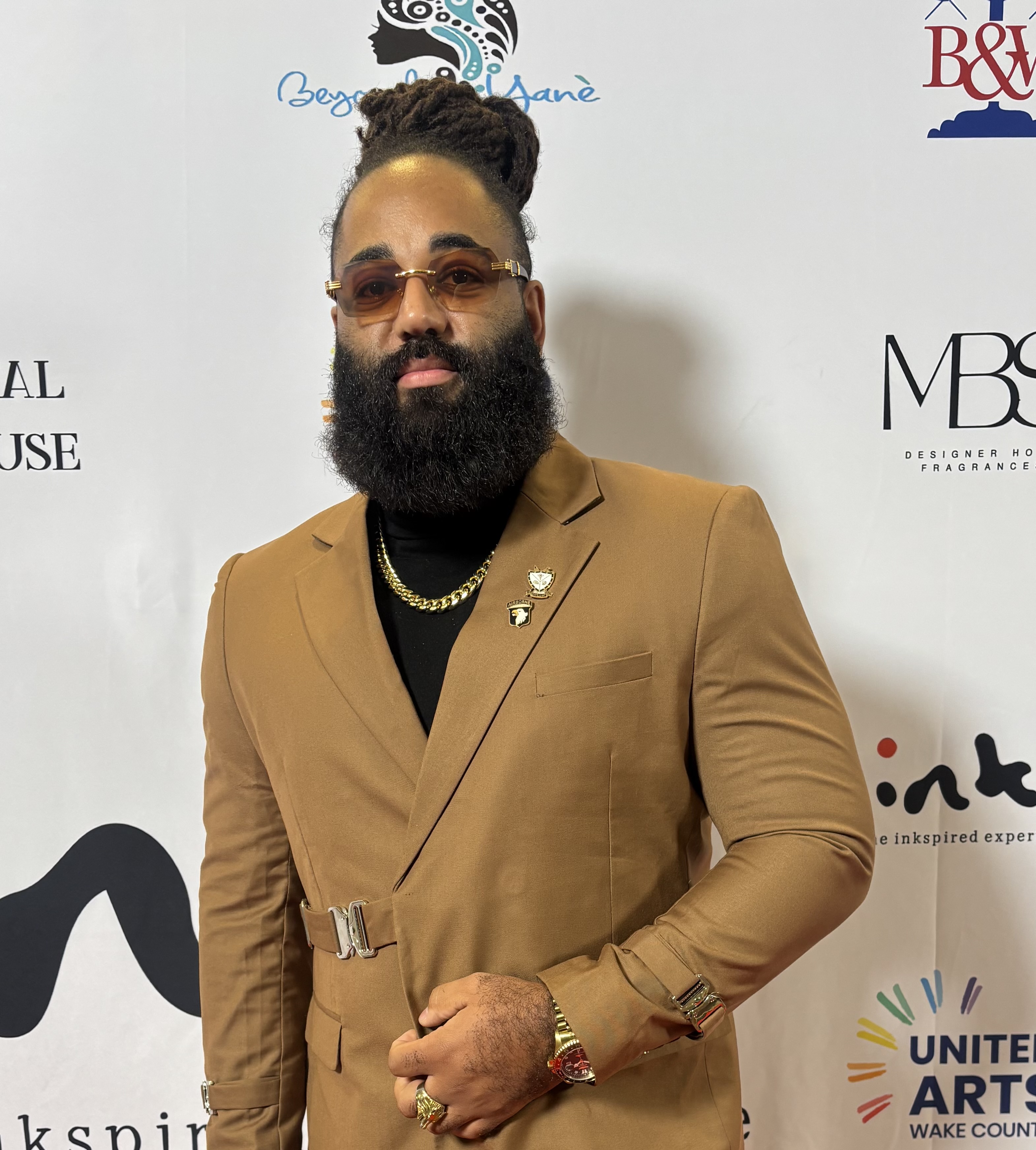 Man wearing a tan suit and black shirt with black hair in a bun and a long black beard. 