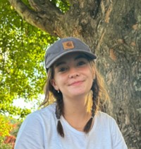 Woman wearing a white top with medium length brown hair in braids wearing a grey hat. 