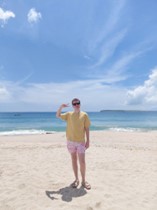 Man on beach with black sunglasses, yellow shirt, and red swim trunks.