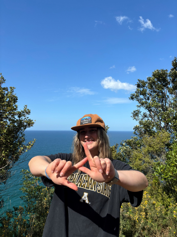 Girl with blonde hair wearing an orange hat wearing an App State shirt holding up a symbol with her hands. Ocean and trees are in the background. 