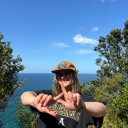 Girl with blonde hair wearing an orange hat wearing an App State shirt holding up a symbol with her hands. Ocean and trees are in the background. 