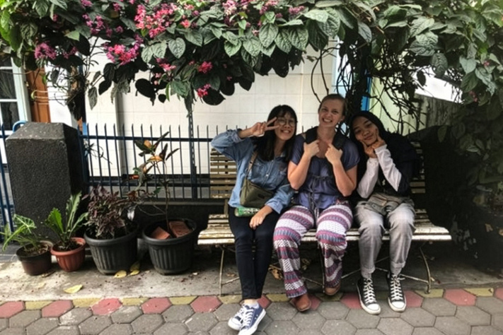 Group of students seated on a bench outdoors, smiling and posing beneath flowering trees.