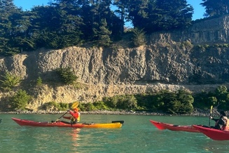Two people kayak on clear turquoise water beneath a rocky cliff lined with dense green trees.