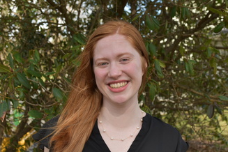 Headshot of woman with red hair smiling with green foilage in the background. 